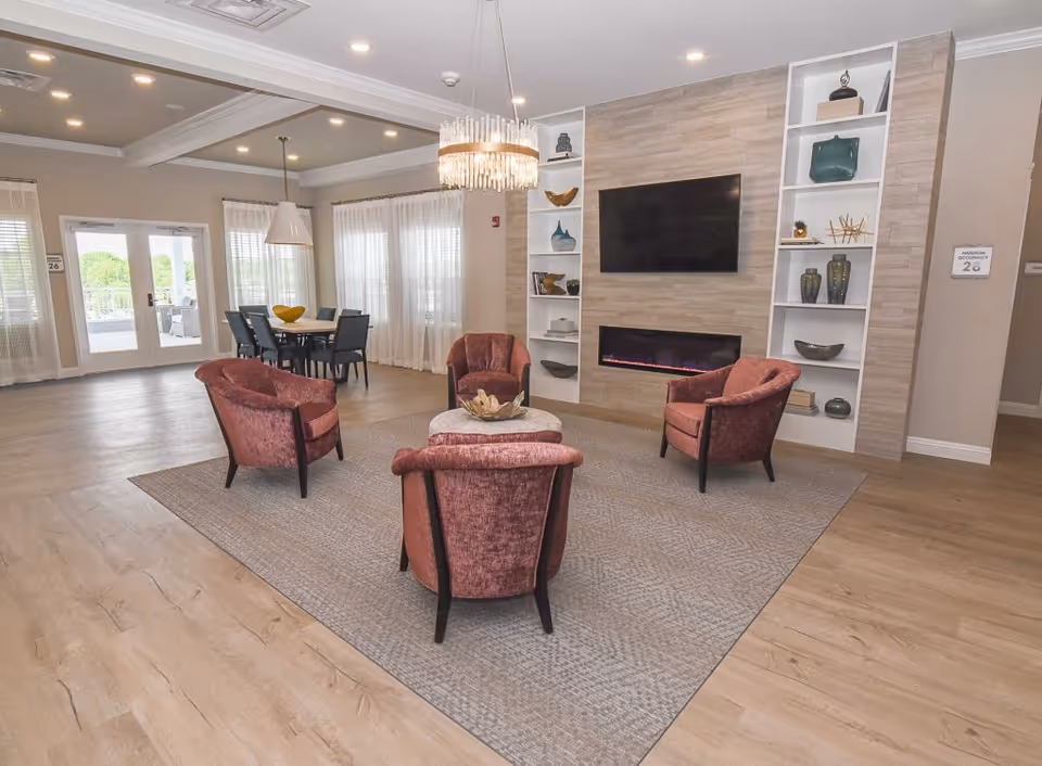 Communal lounge with four pink upholstered chairs around a small table on a rug, a wall-mounted TV and linear fireplace flanked by shelves, and a dining area by windows.