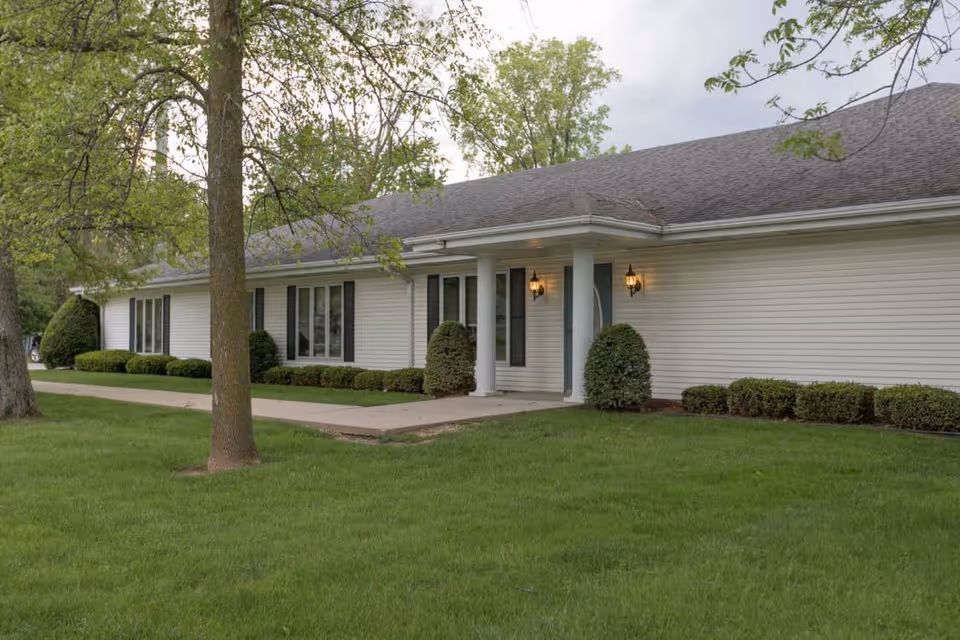 Exterior view of Cottonwood Manor Assisted Living, a single-story building with white siding, a gray shingled roof, and a covered entrance supported by two white columns. The building is surrounded by green grass, trimmed bushes, and trees.