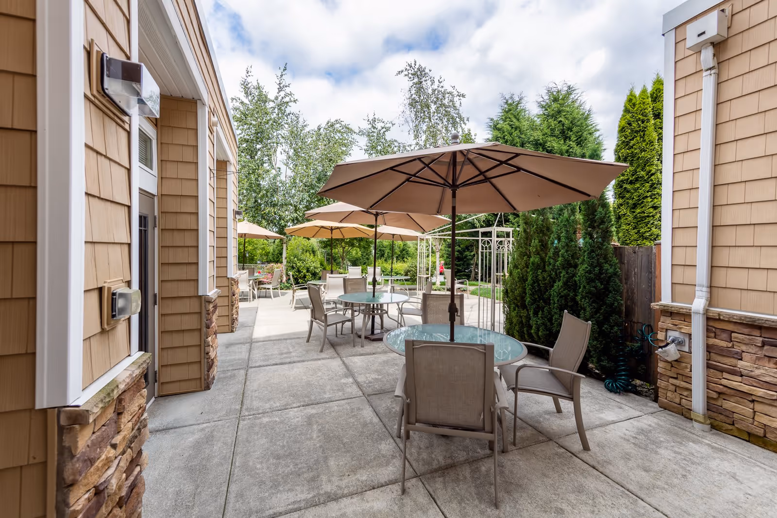 Outdoor patio area at Woodland Terrace with multiple round glass tables, each shaded by large beige umbrellas. Several chairs are arranged around the tables. The patio is bordered by tan siding walls with stone accents and surrounded by green trees and shrubs under a partly cloudy sky.