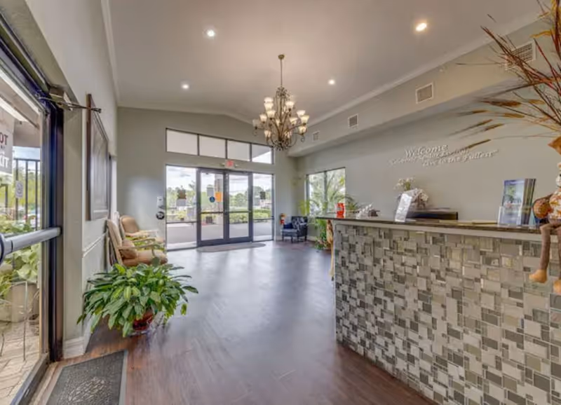 Reception area of Regency Park Assisted Living & Memory Care with a tiled front desk, chandelier lighting, seating area with chairs, large windows and glass doors letting in natural light, and indoor plants.