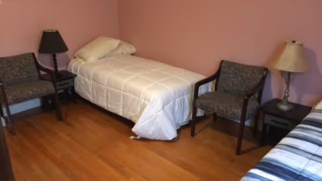 A small bedroom with two single beds, each with a pillow and bedding. There are two upholstered chairs with wooden arms, one on each side of the room. Two bedside tables with lamps are positioned next to each bed. The walls are painted pink and the floor is wooden.