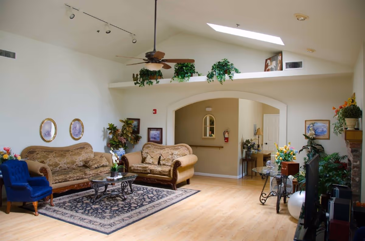 A bright and spacious living room with two beige patterned sofas, a blue armchair, a glass coffee table on a patterned rug, and various plants and framed pictures on the walls. The room features a ceiling fan, skylight, and an arched doorway leading to another area.