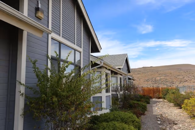Exterior view of a gray building with white trim, featuring multiple windows and a pathway lined with bushes and small trees. The sky is clear with some clouds, and there is a dry, hilly landscape in the background.