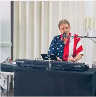 An elderly man wearing a shirt with an American flag design is playing a Yamaha MODX7 keyboard and singing into a microphone in an indoor setting with beige curtains in the background.