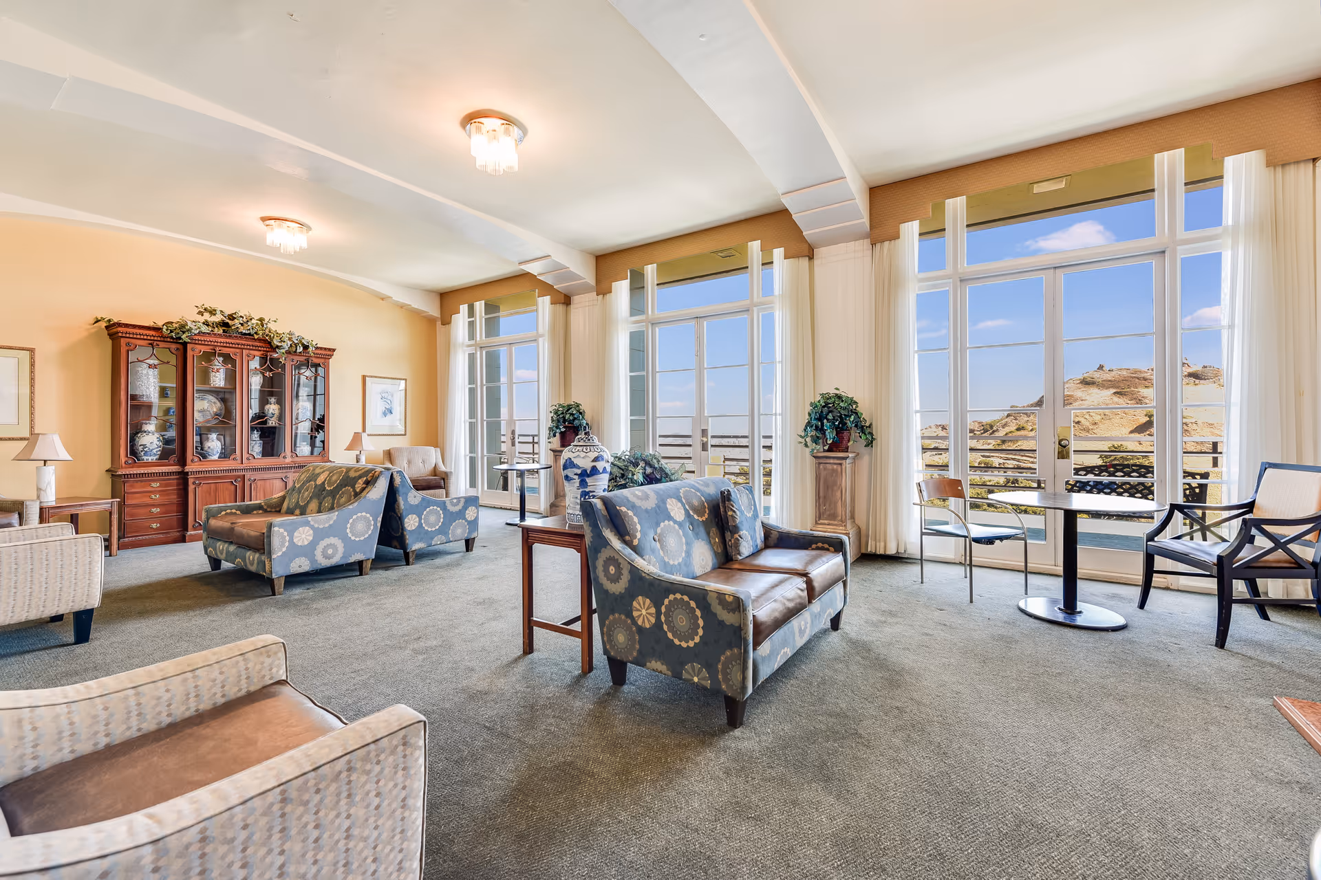 A spacious senior living lounge with upholstered sofas and chairs, a china cabinet, and large floor-to-ceiling windows overlooking a hillside view.