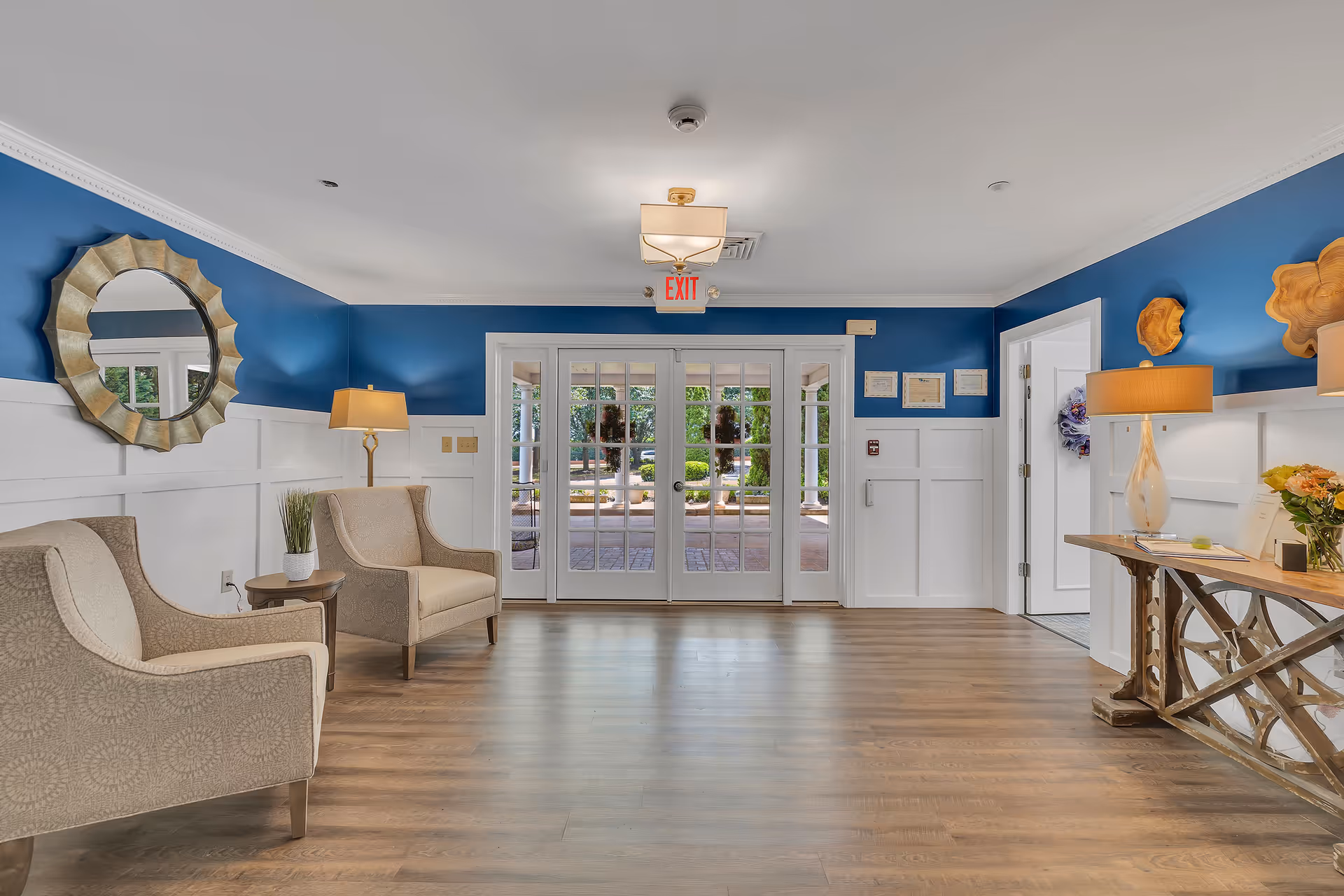 A bright and welcoming interior room with blue and white walls, two beige armchairs with a small table and lamp between them on the left, a decorative round mirror on the wall, and a wooden console table with a lamp and flowers on the right. Large glass double doors lead outside to a garden area with greenery visible through the doors.