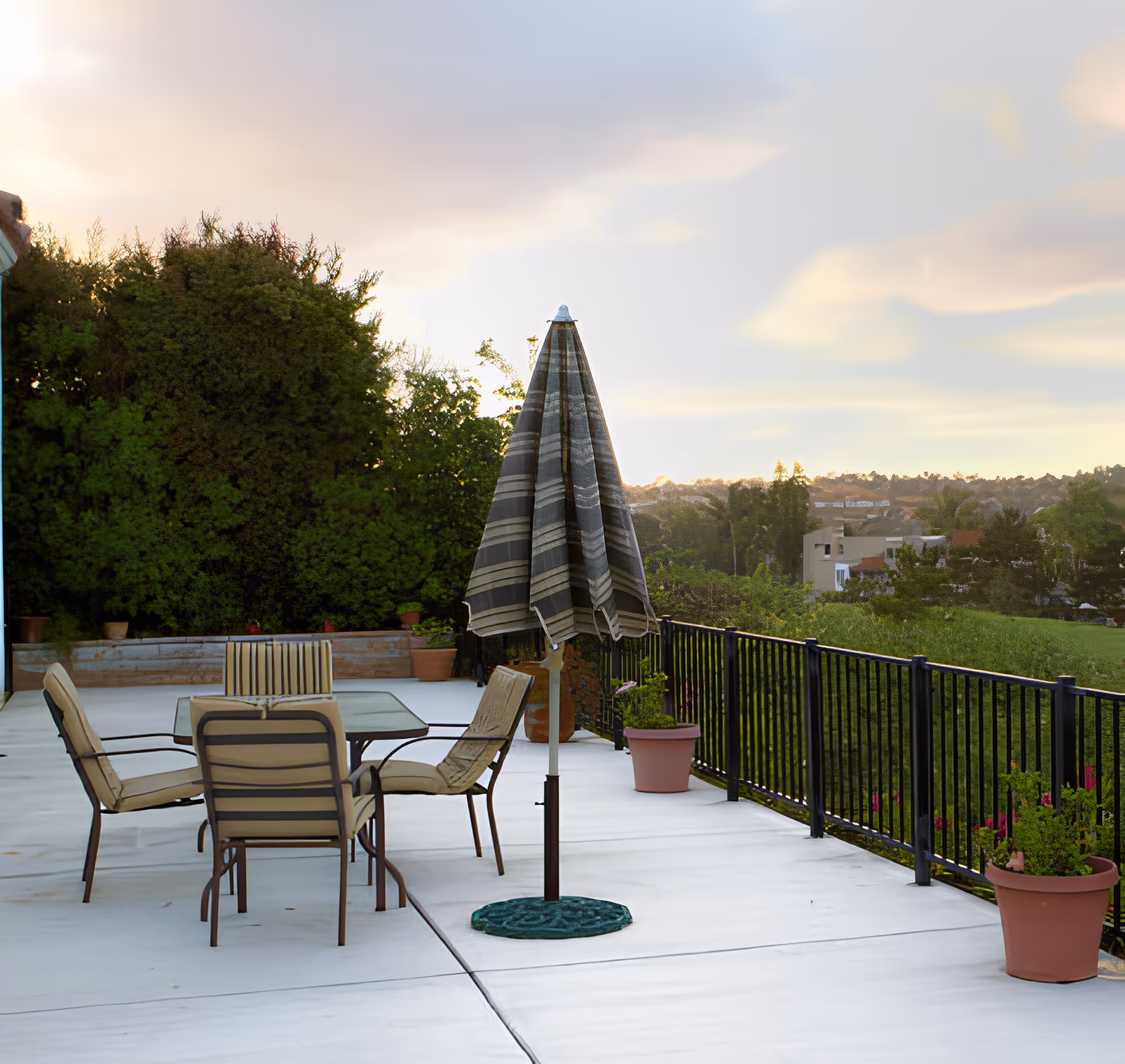 Outdoor patio with a table, four chairs, and a closed striped umbrella overlooking greenery and distant houses.
