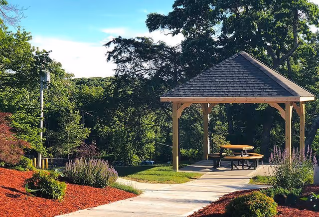 A wooden gazebo with a shingled roof situated in a landscaped outdoor area with a concrete pathway leading to it. The gazebo contains a round picnic table with attached benches. Surrounding the pathway are mulched garden beds with various shrubs and flowering plants, and tall trees in the background under a clear blue sky.