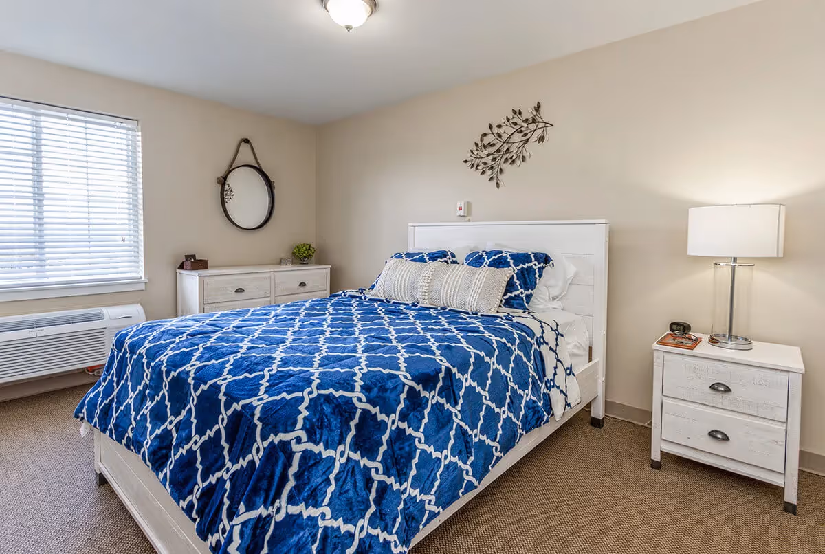 Bright bedroom with a blue-patterned bedspread, white wooden bed and nightstands, a lamp, dresser and window with blinds.