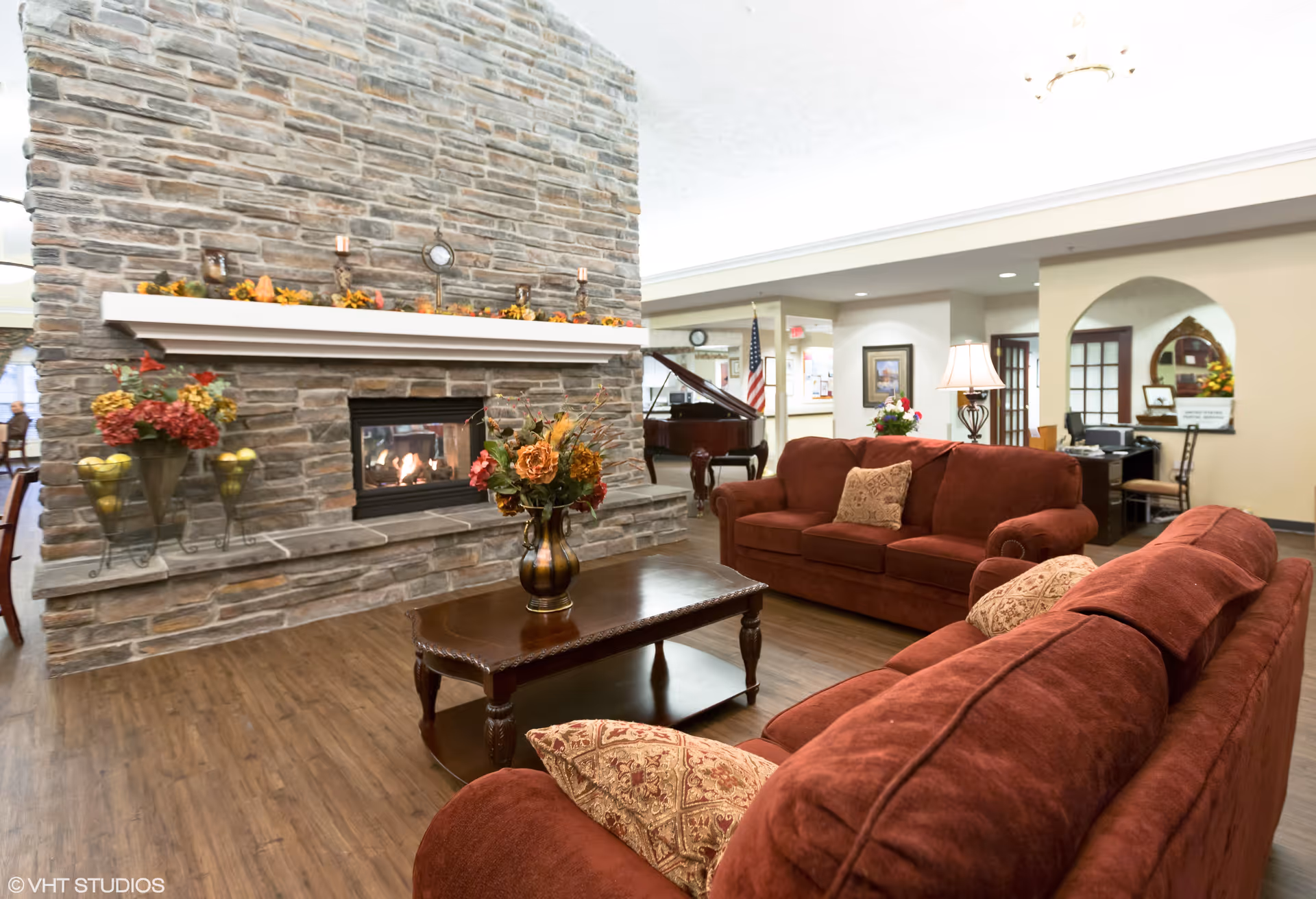 A cozy living room area with two red sofas and a wooden coffee table with a floral arrangement. There is a large stone fireplace with a mantle decorated with candles and autumn-themed decorations. In the background, a grand piano and an American flag are visible, along with a desk and lamp in a well-lit, open interior space.