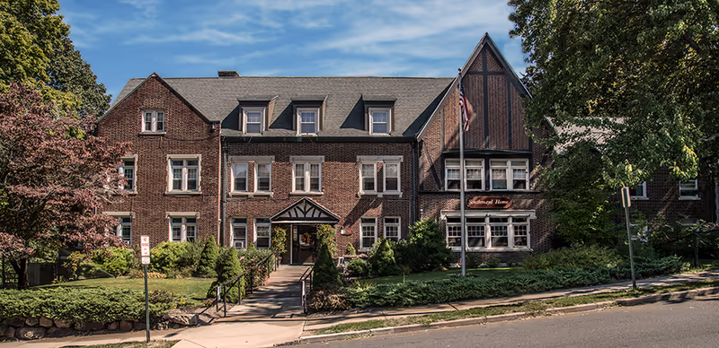 Exterior view of a large brick building with multiple windows and a steep roof, surrounded by trees and greenery. A sidewalk and a ramp lead to the main entrance, and an American flag is displayed on a flagpole near the entrance. The building has a sign that reads 'Southmayd Home'.