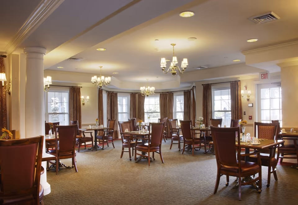 A spacious dining room with multiple wooden tables and chairs arranged neatly. Each table has small flower vases and place settings. The room is well-lit with chandeliers and wall sconces, and large windows with brown curtains allow natural light to enter.