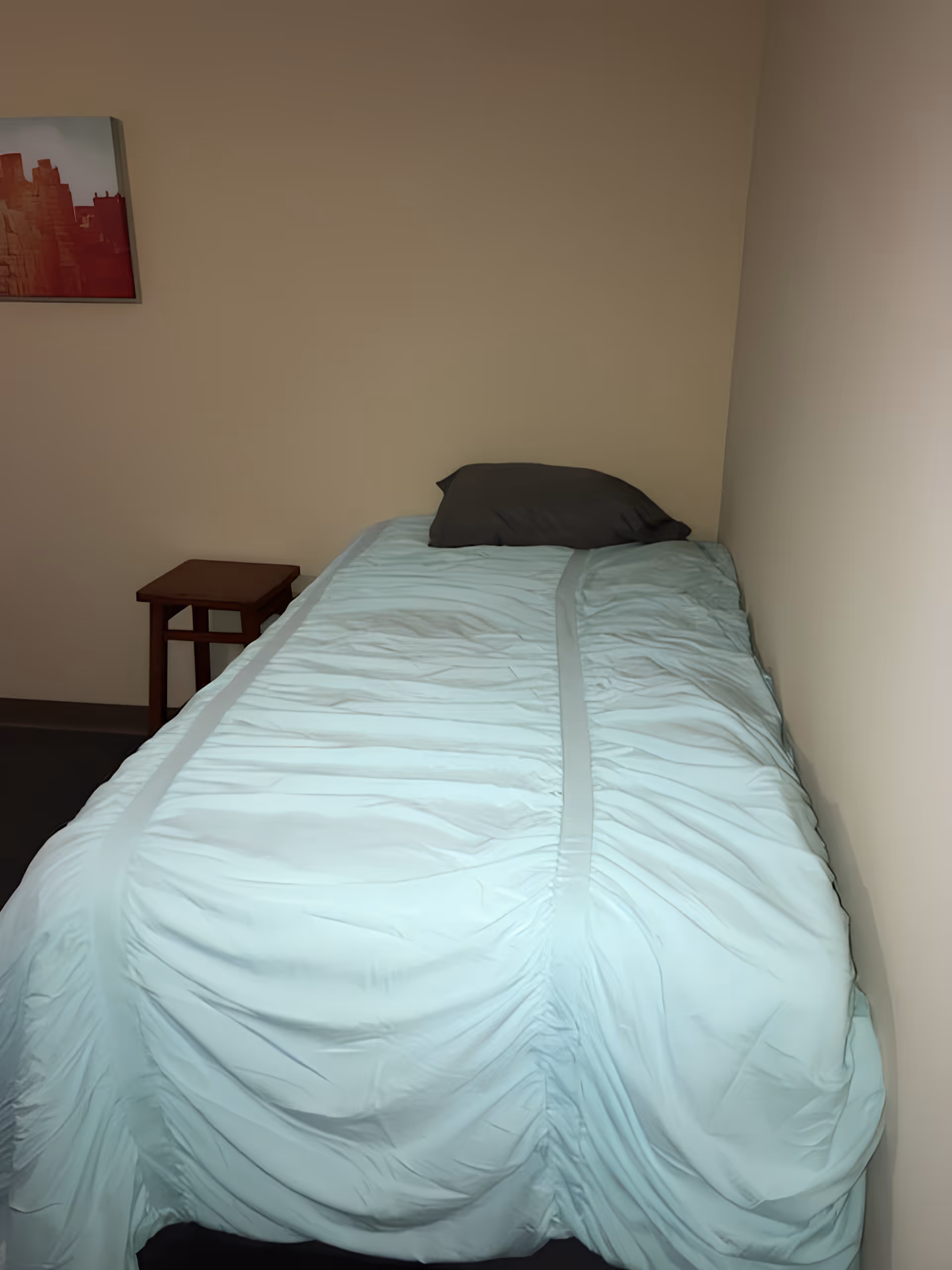 A small bedroom with a single bed against the wall, covered with a light blue bedspread and a dark pillow. There is a small wooden side table next to the bed and a framed picture hanging on the beige wall.