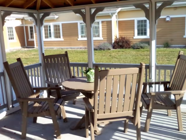 Covered outdoor gazebo with a round table and four wooden chairs overlooking a grassy courtyard and building.