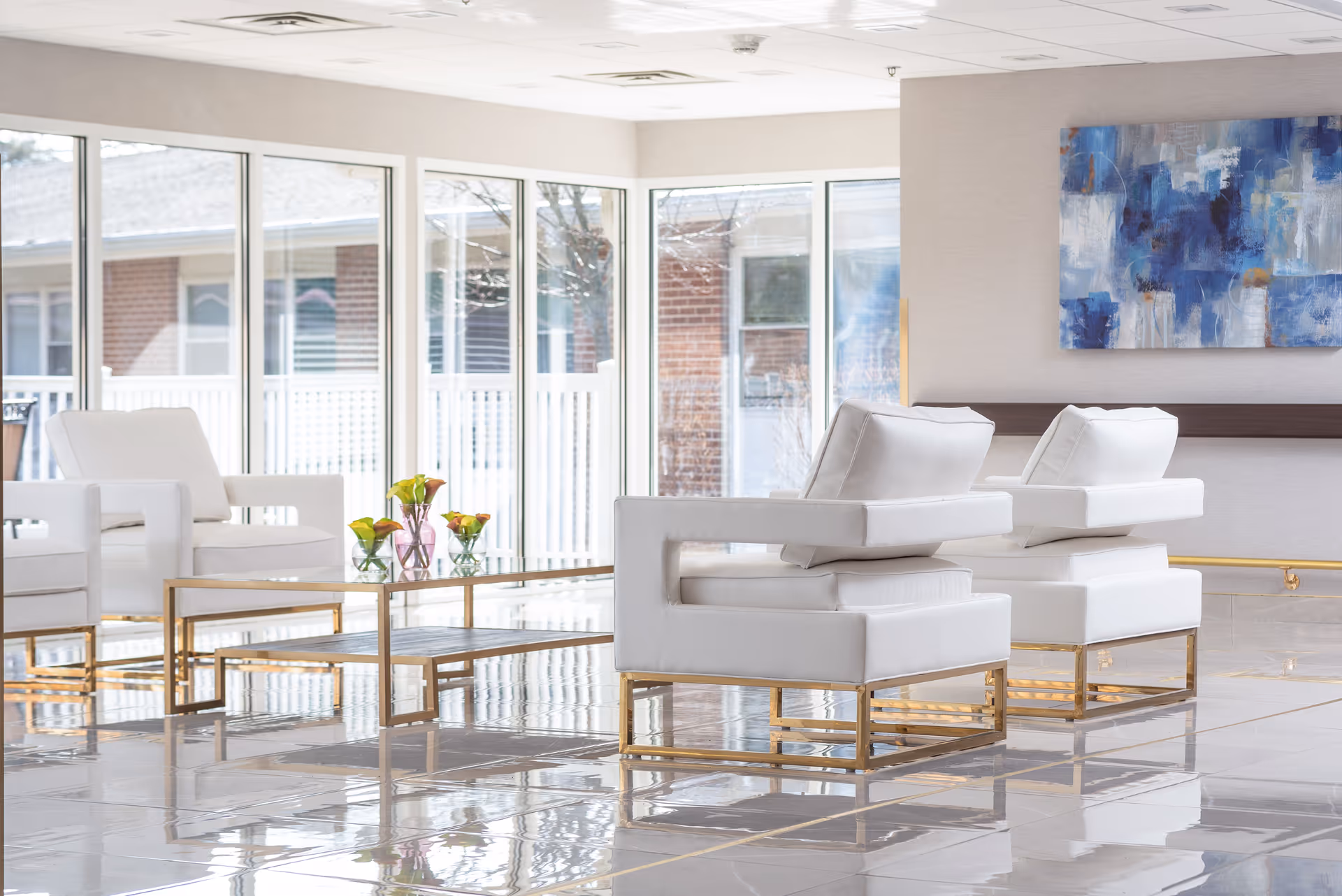 Bright and modern living room area with white cushioned chairs featuring gold metal frames, a glass coffee table with gold legs, and large windows letting in natural light. A blue abstract painting hangs on the wall.