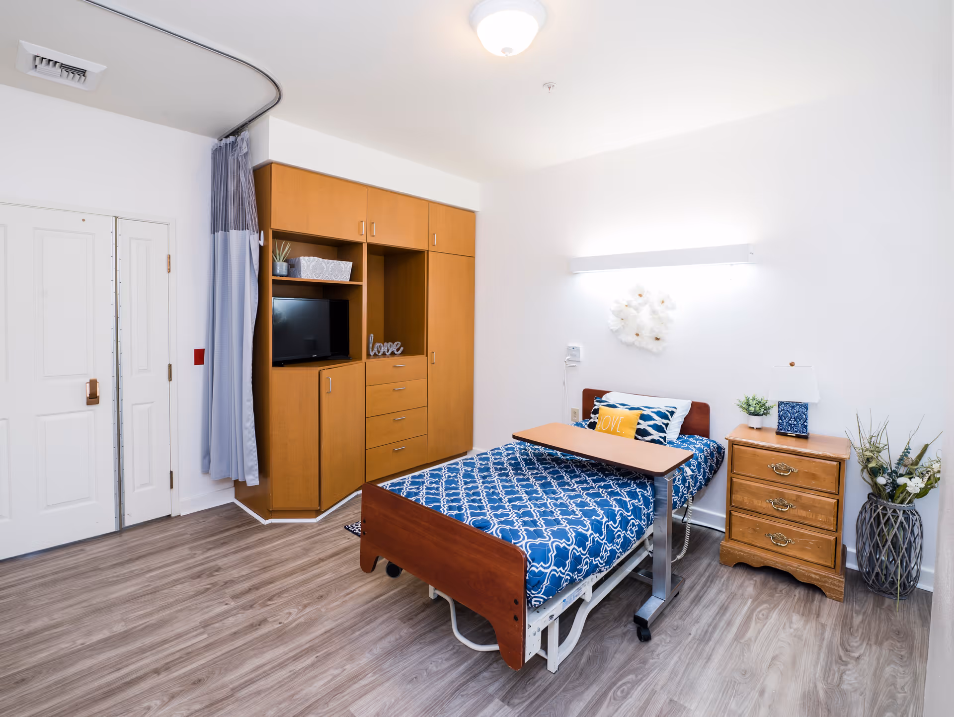 A clean and bright senior living bedroom with a single bed featuring a blue patterned bedspread and a yellow pillow with the word 'LOVE'. The room has wooden flooring, a wooden bedside table with a lamp and plant, a large wooden cabinet with shelves and drawers, and a TV. A privacy curtain is partially drawn near the door.