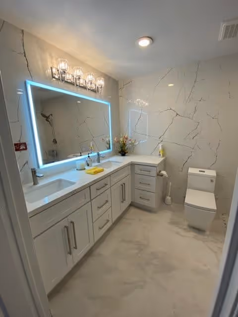 Modern bathroom with white marble walls and floor, a large illuminated mirror above a double sink vanity with multiple drawers and cabinets, a toilet, and a shower area visible in the mirror reflection.