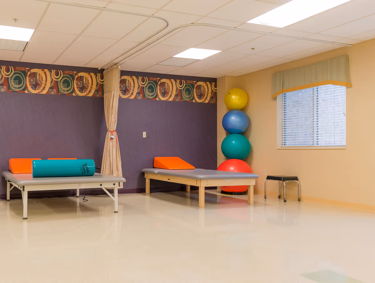 A physical therapy room with two padded therapy tables, each with an orange wedge cushion. There is a stack of four large colorful exercise balls in the corner next to a window with blinds and a valance. A small black stool is near the window. The walls are beige and purple with a decorative border near the ceiling.