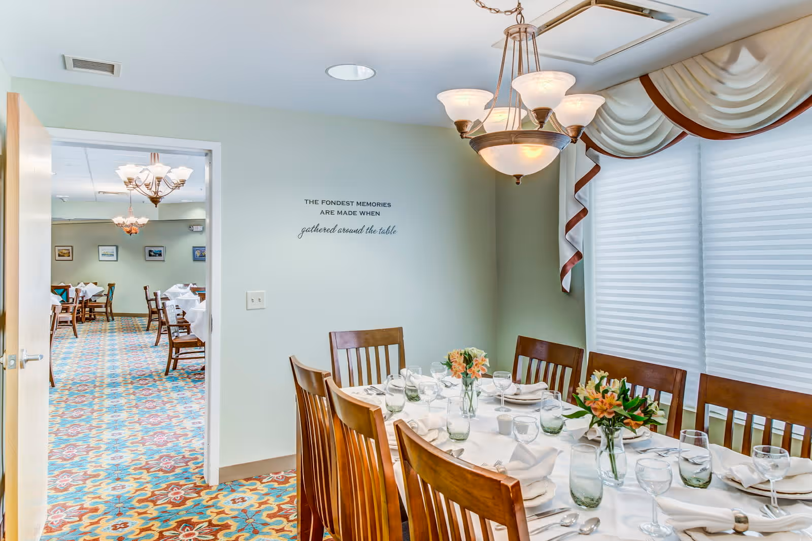 A dining room with a table set for a meal, featuring wooden chairs, white tablecloth, glassware, napkins, and floral centerpieces. The room has a chandelier light fixture and large windows with white blinds and decorative valances. Through an open door, another dining area with multiple tables and chairs is visible. On the wall, there is a quote that reads, 'THE FONDEST MEMORIES ARE MADE WHEN gathered around the table.'