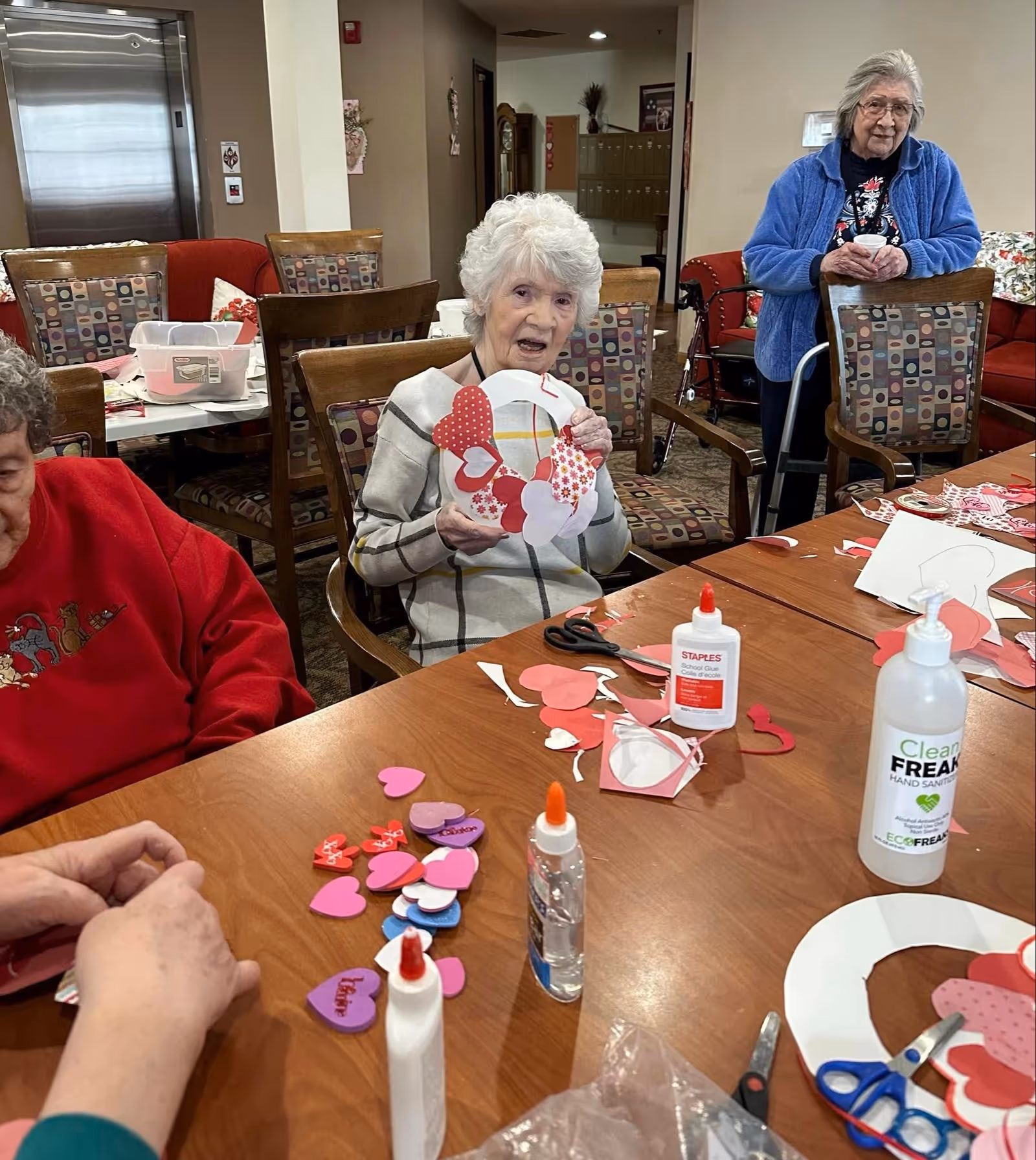 Three elderly women sitting and standing around a table in a communal room, engaging in a craft activity with paper hearts, glue, scissors, and hand sanitizer visible on the table. One woman is holding a heart-themed wreath, while another is working with paper hearts. The room has patterned chairs and a red couch in the background.