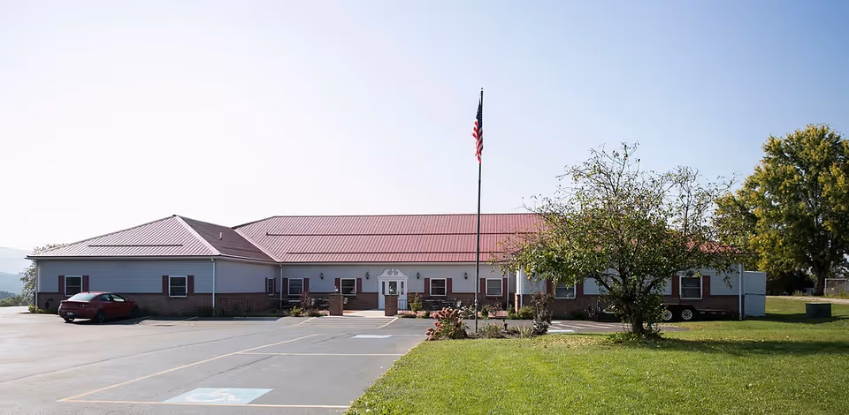 Single-story building with a red roof and white siding, surrounded by a parking lot and green lawn. An American flag is flying on a flagpole in front of the building, with a tree to the right side. The sky is clear and blue.