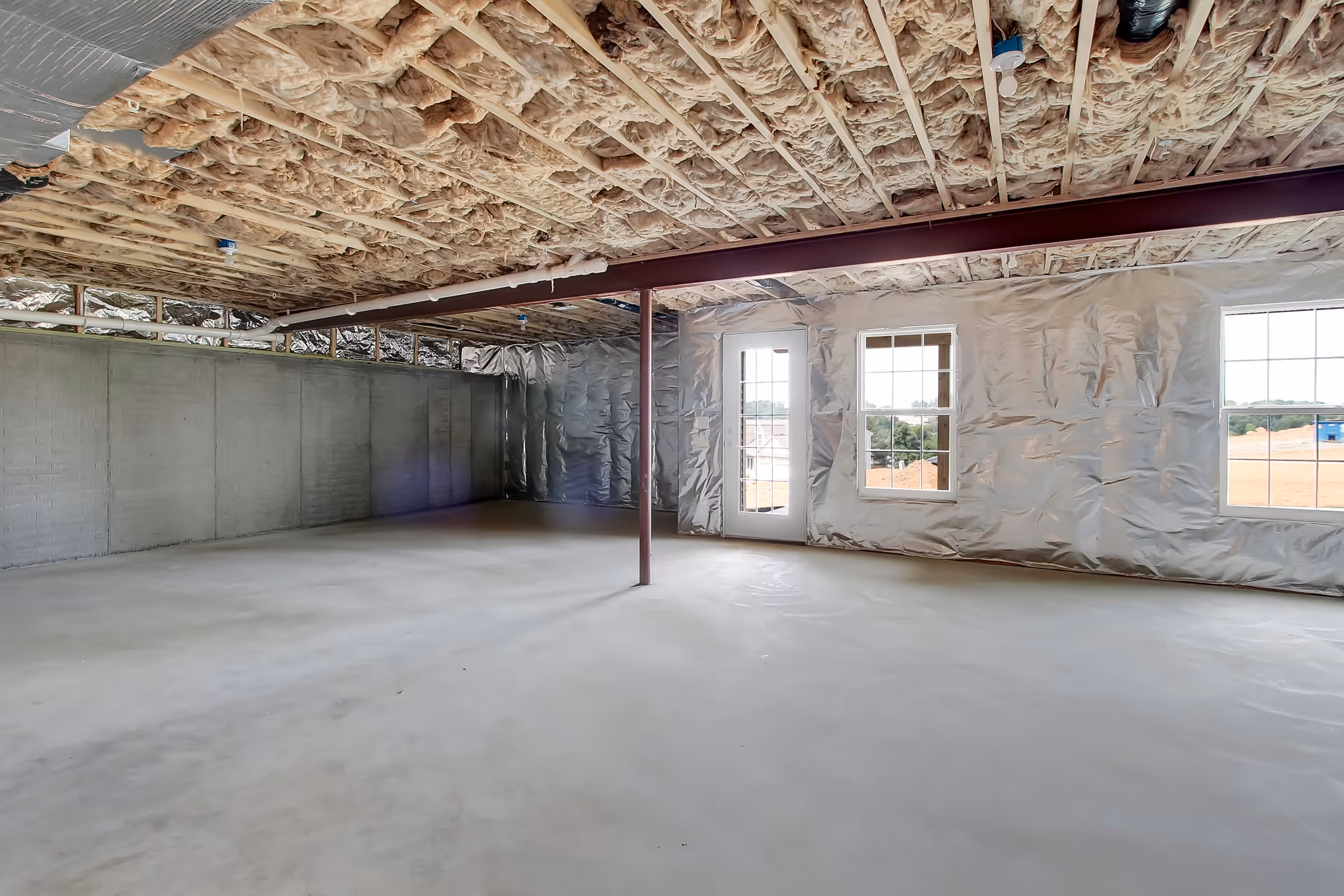An unfinished basement or large room with exposed insulation on the ceiling and walls, concrete floor, a metal support beam in the center, and three windows including a glass door letting in natural light.