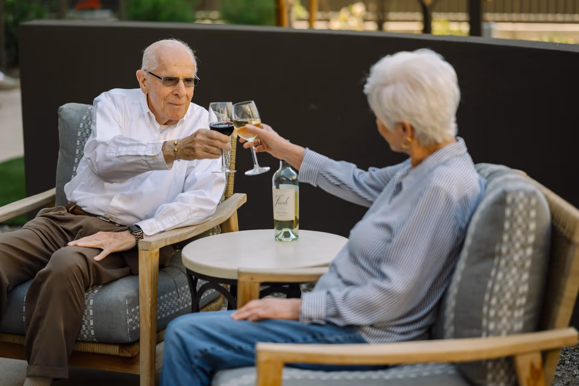 An elderly man and woman sitting outdoors on cushioned chairs around a small round table, clinking wine glasses in a toast. There is a bottle of white wine on the table. The man is wearing a white shirt and brown pants, and the woman is wearing a striped shirt and blue jeans.