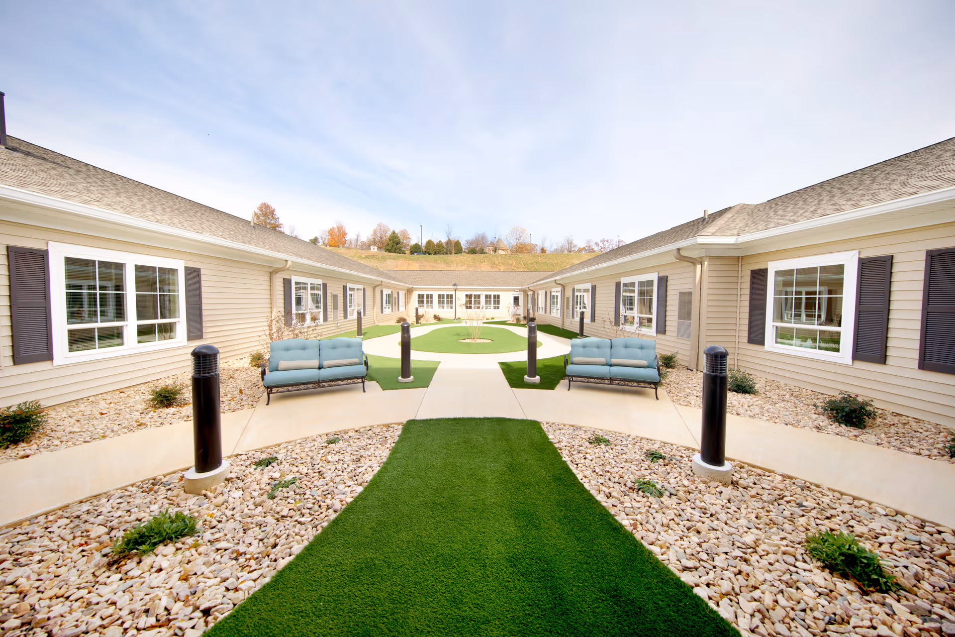 Outdoor courtyard area of a senior living facility with beige siding buildings on both sides, multiple windows with dark shutters, two blue cushioned benches facing each other, pathways, green artificial turf, and decorative rocks along the edges. The sky is clear with a few clouds.