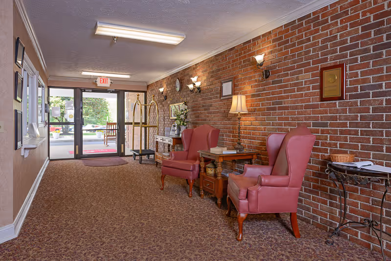 Interior view of a senior living facility lobby with two red upholstered armchairs, a wooden side table with a lamp and books, a brick wall with framed certificates and a clock, a luggage cart, and glass double doors leading outside.