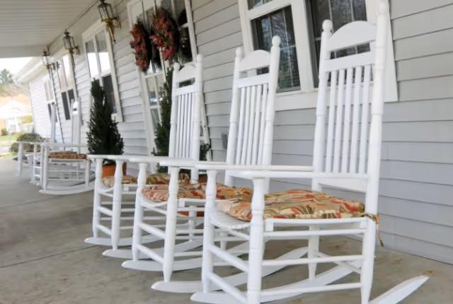 A row of white wooden rocking chairs with floral cushions on a covered porch outside a building with gray siding. There are decorative wreaths on the windows and potted plants along the porch.