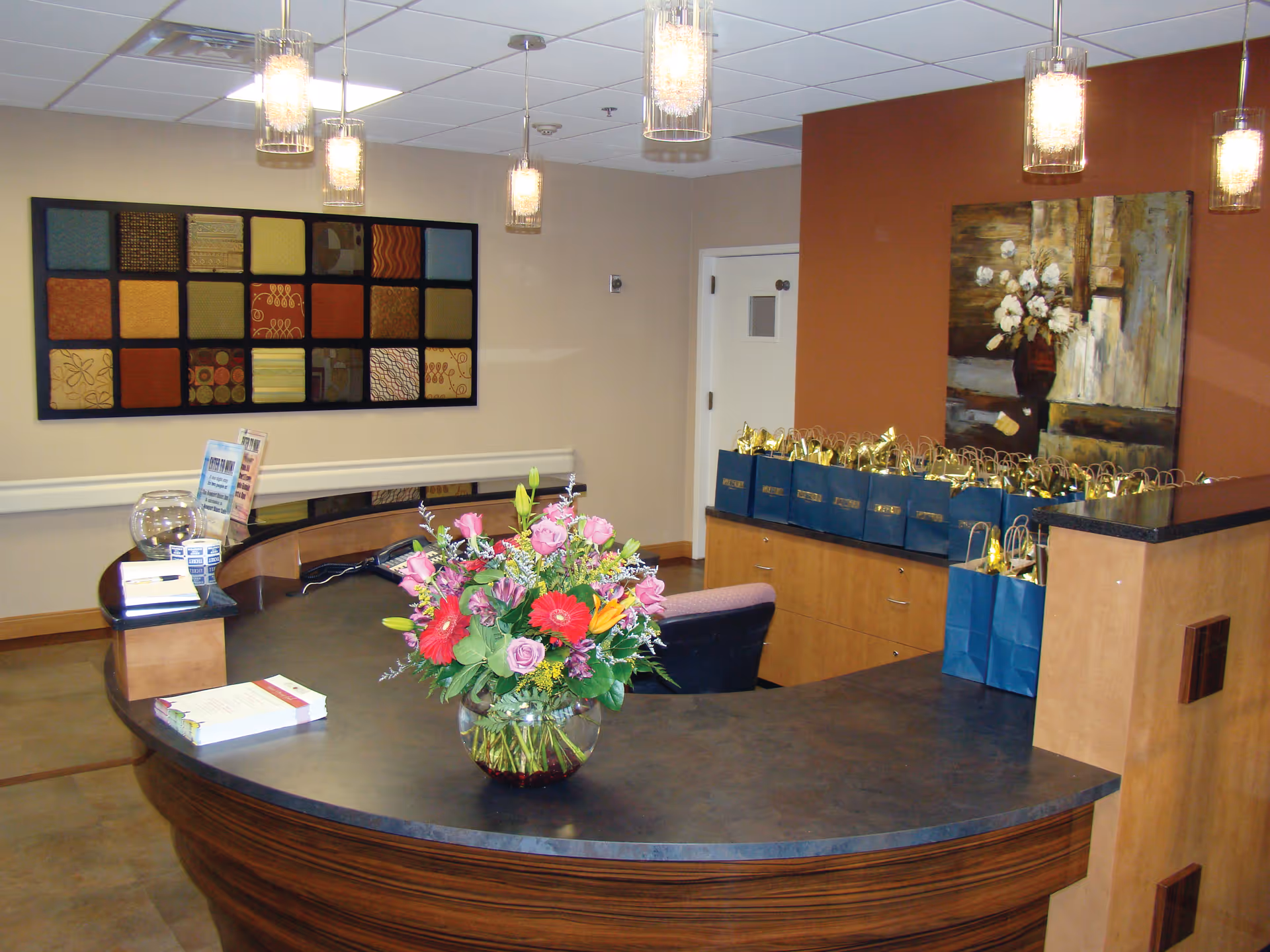 Reception desk area with a curved dark countertop and wooden base, decorated with a large bouquet of colorful flowers in a glass vase. Behind the desk are multiple blue gift bags with gold tissue paper on a wooden cabinet. The walls are painted beige and burnt orange, adorned with a large abstract painting and a decorative wall panel with various fabric squares. Several pendant lights hang from the ceiling.