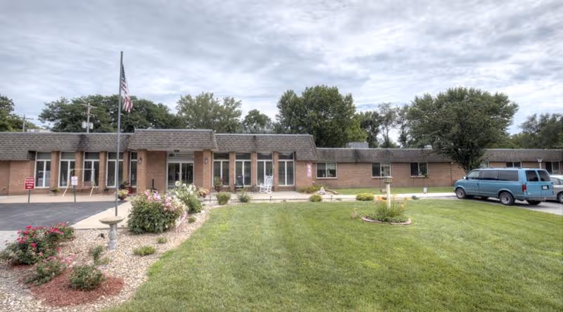 Front exterior view of Wathena Healthcare and Rehabilitation Center, a single-story brick building with multiple windows, an American flag on a flagpole, a well-maintained lawn, flower beds, and a blue van parked on the right side.