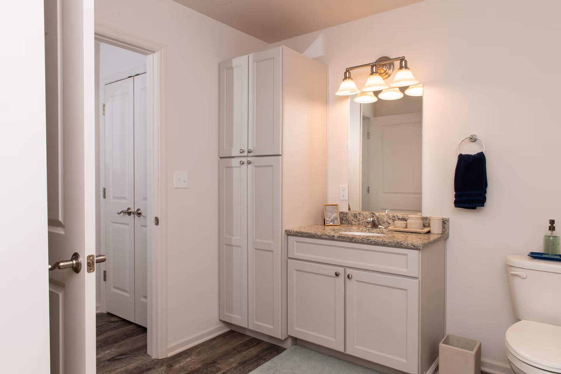 A clean and modern bathroom featuring a white vanity with granite countertop, a large mirror with four light fixtures above it, a tall white cabinet for storage, a toilet with a soap dispenser on top, a towel ring with a dark blue towel, and wood-style flooring.