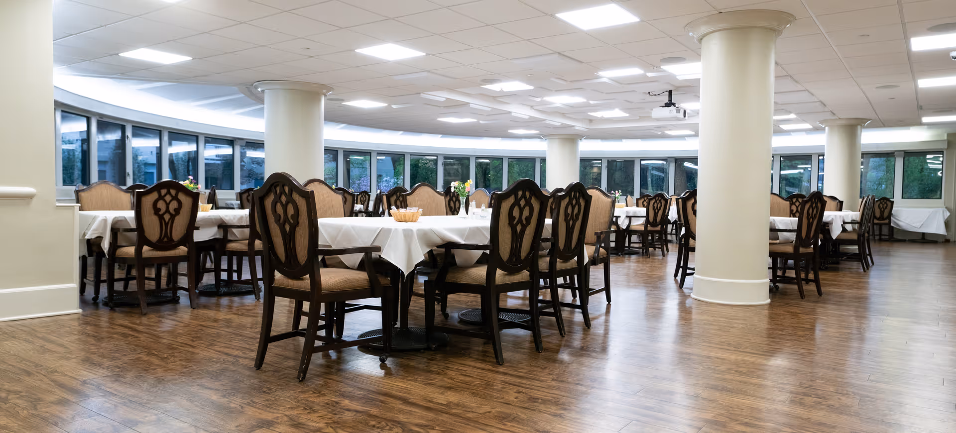 A spacious dining room with multiple tables covered in white tablecloths and surrounded by wooden chairs with cushioned seats and decorative backs. The room has large windows along the curved wall, allowing natural light to enter, and a wooden floor with white columns supporting the ceiling.
