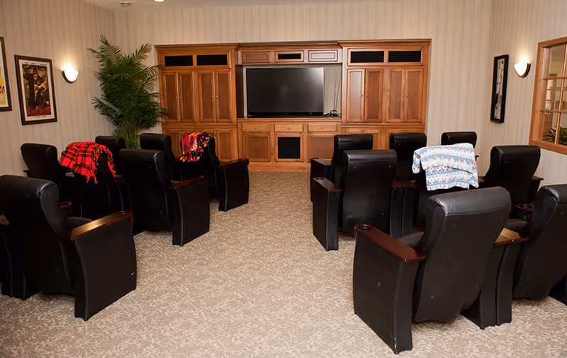 A cozy common room with black leather recliner chairs arranged in rows facing a large flat-screen TV mounted in a wooden entertainment center. Some chairs have colorful blankets draped over them. The room has beige carpet, light-colored wallpaper, framed artwork on the walls, and a potted plant in the corner.