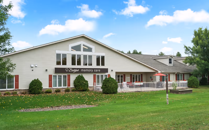 Exterior view of a single-story assisted living and memory care facility building with beige siding, red shutters, and a sign that reads 'Bickford memory care'. The building is surrounded by green grass, bushes, and trees under a partly cloudy blue sky.