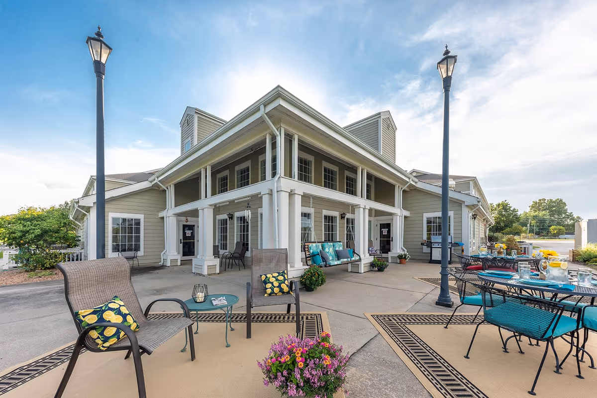 Outdoor patio area at Fort Scott Presbyterian Village featuring seating arrangements with chairs and tables, decorative cushions, potted plants, and two tall street lamps, adjacent to a beige building with white trim under a partly cloudy sky.