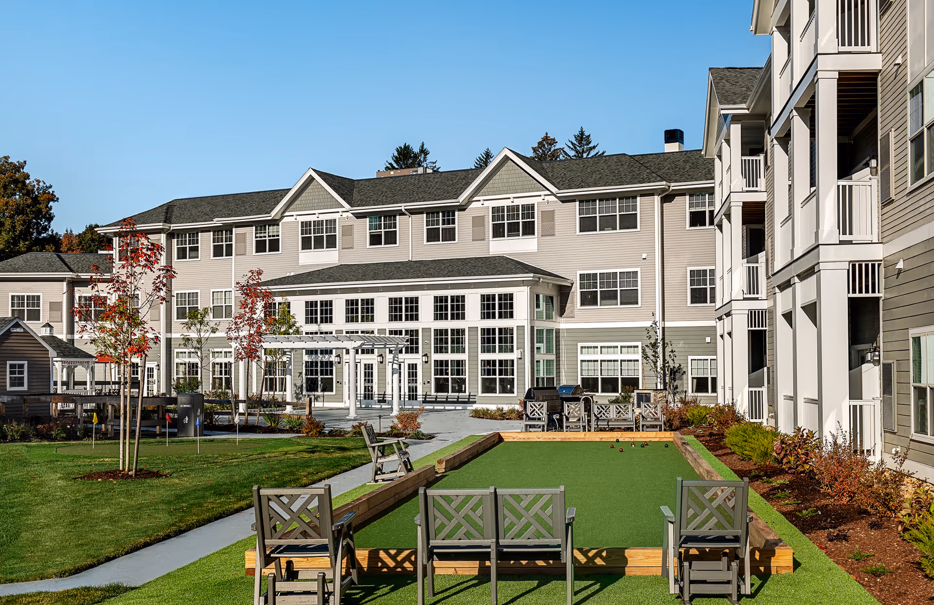 Outdoor view of a retirement community building with a bocce ball court in the foreground, surrounded by chairs and greenery under a clear blue sky.