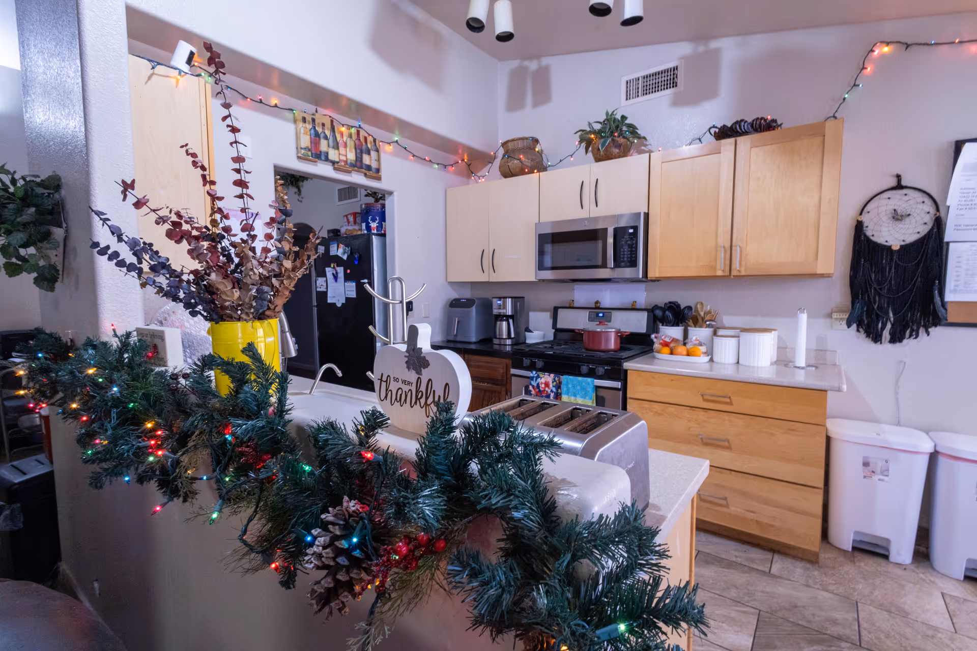 Interior view of a kitchen decorated with holiday lights and greenery. The kitchen features light wood cabinets, a microwave, a stove with a red pot, a coffee maker, and a toaster. There is a yellow vase with dried flowers on the counter, a decorative sign that says 'so very thankful,' and a dreamcatcher hanging on the wall. Two white trash bins are visible on the right side.