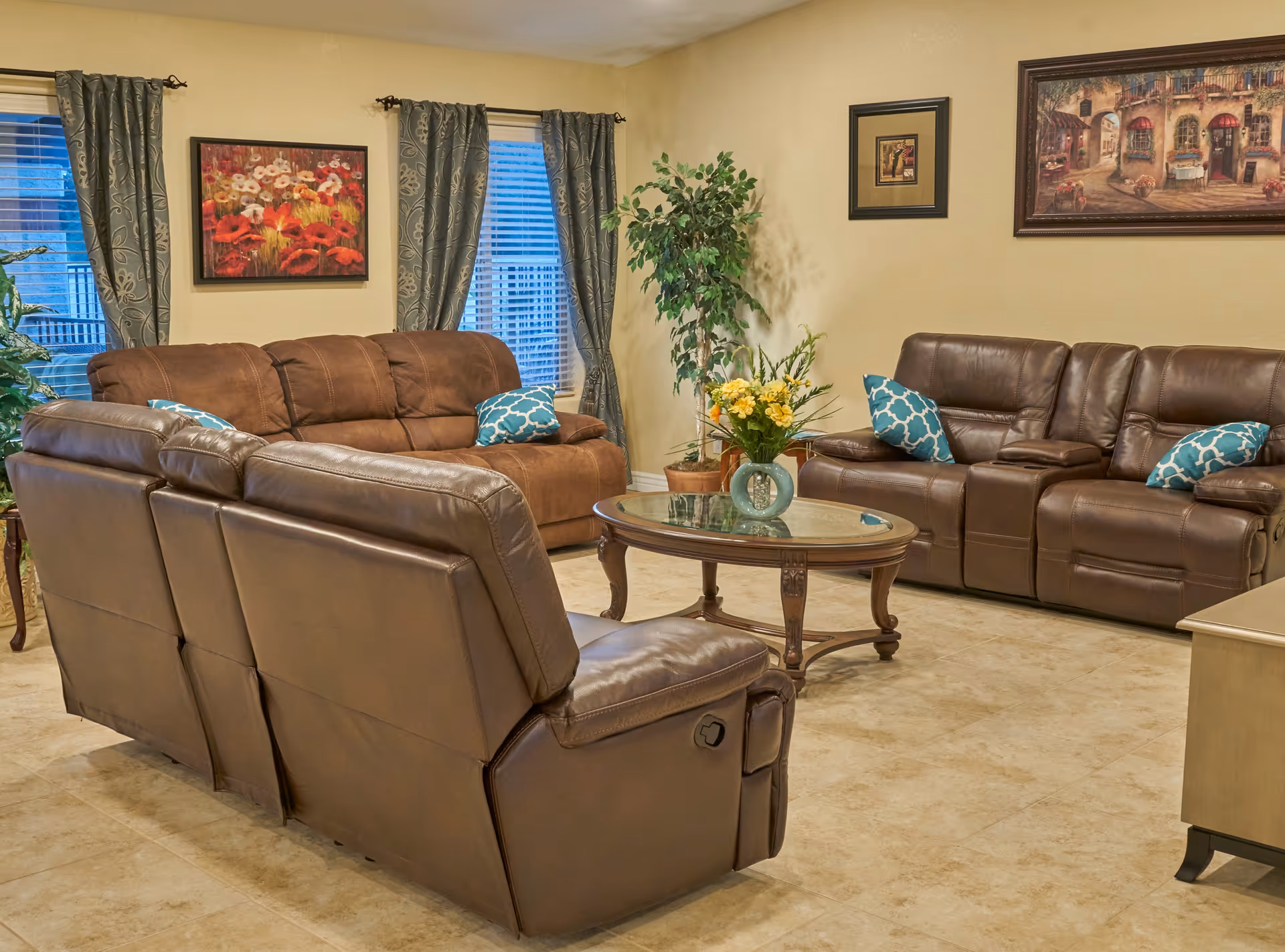 A cozy living room with two brown leather sofas and one brown fabric sofa arranged around a glass-top wooden coffee table with a vase of yellow flowers. The room has beige walls, tiled floors, two windows with patterned curtains, and framed artwork on the walls. There are also two potted plants adding greenery to the space.