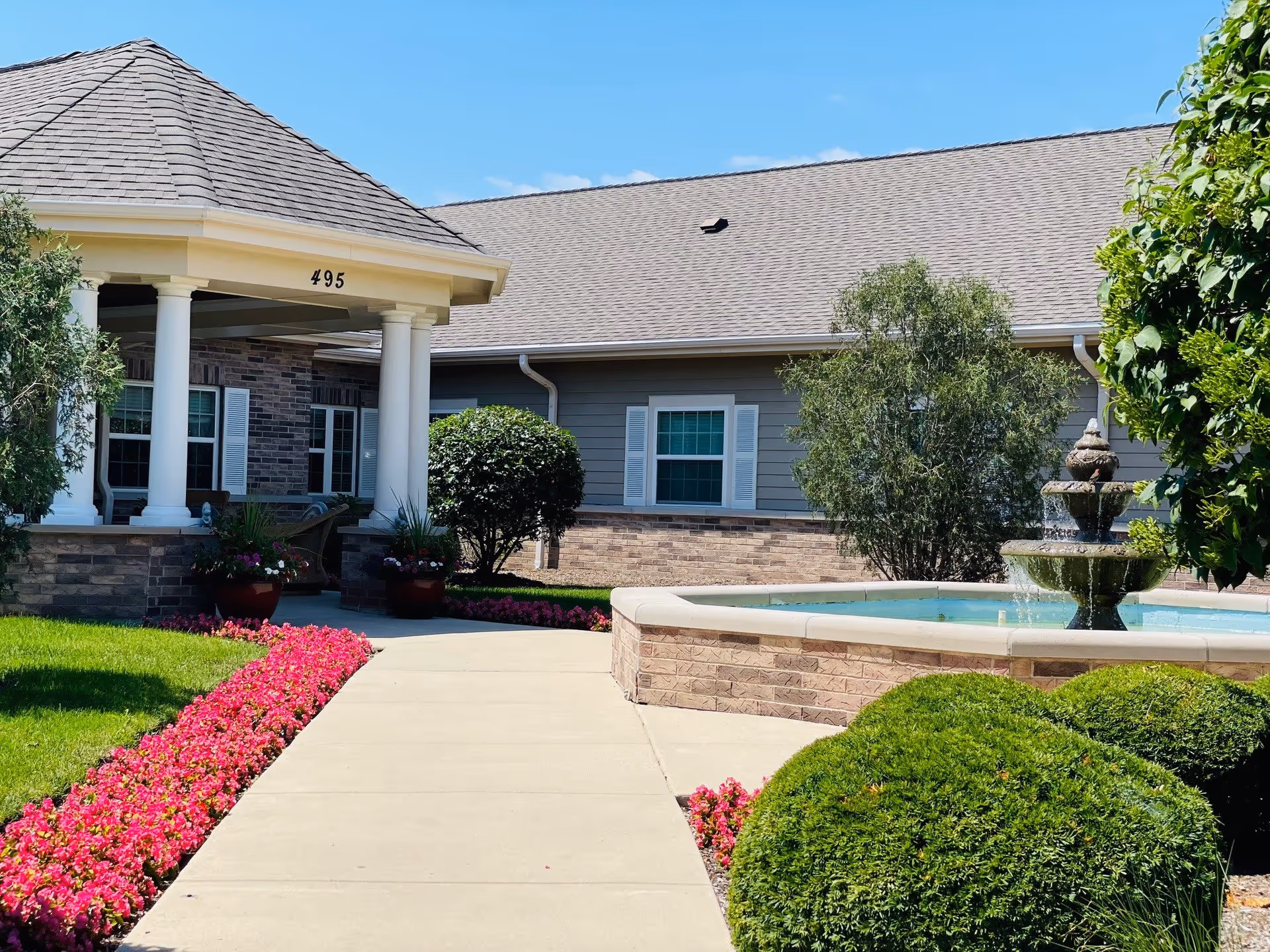 Outdoor view of a senior living facility entrance with a covered porch supported by white columns, a walkway lined with vibrant pink flowers, neatly trimmed bushes, and a decorative water fountain. The building has a gray exterior with white window shutters under a clear blue sky.