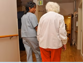 A healthcare worker in gray scrubs walking down a hallway with an elderly woman wearing a white shirt and red pants in a senior care facility.