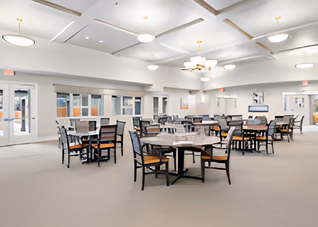 Spacious senior living dining room with multiple round tables and chairs under ceiling lights.