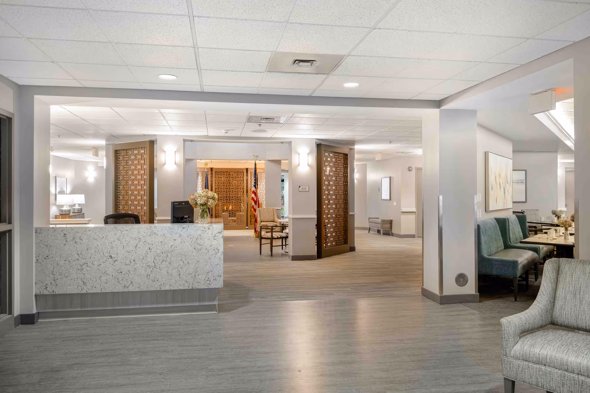 A spacious and well-lit senior living community interior featuring a reception desk with a computer and a vase of flowers. The area has modern decor with light-colored walls, wood-patterned flooring, and decorative wooden panels. To the right, there is a seating area with green upholstered chairs and a table, and in the background, an American flag is visible near a pair of chairs.