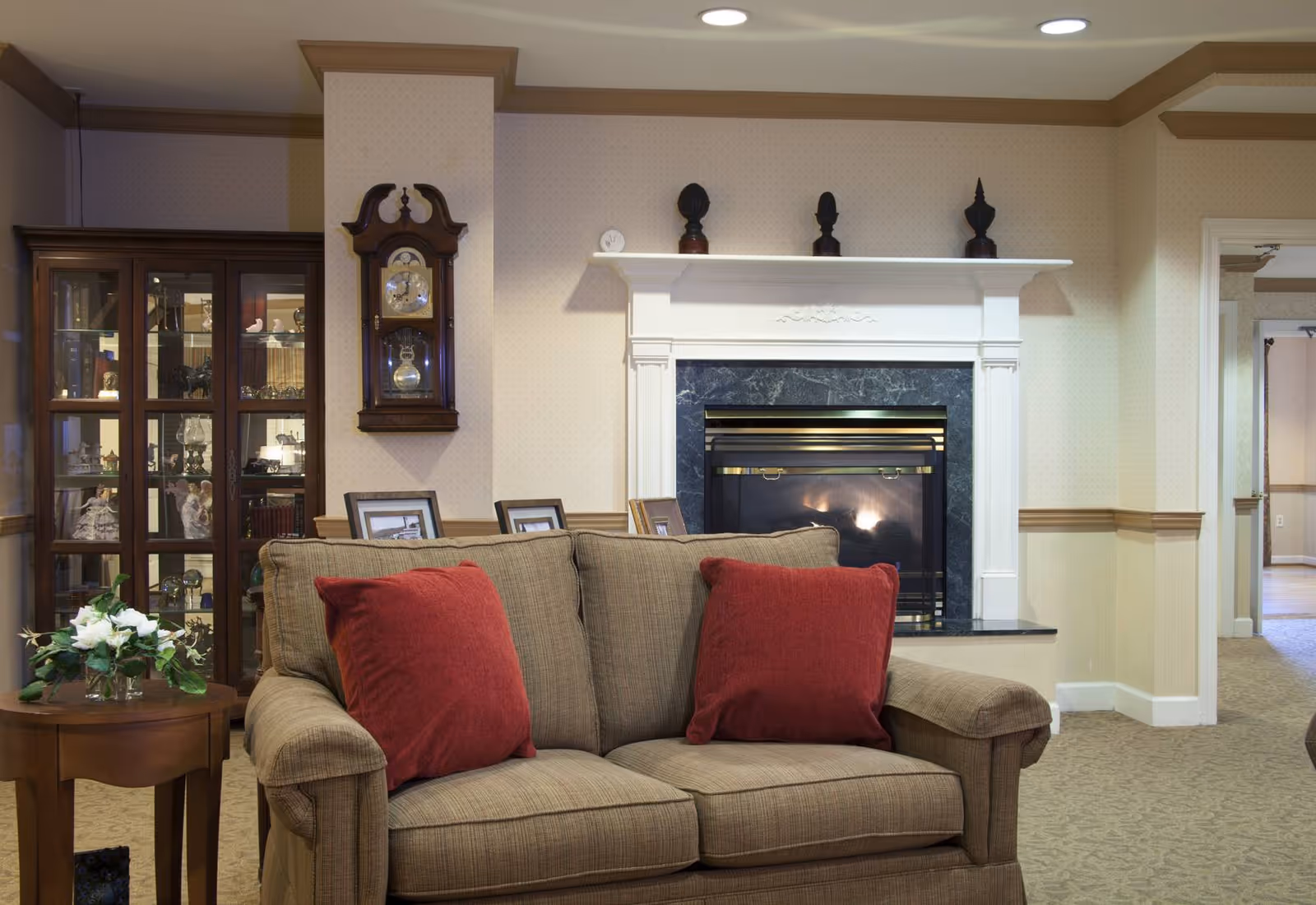 Beige loveseat with red throw pillows placed in front of a marble fireplace and a glass display cabinet in a cozy living room.