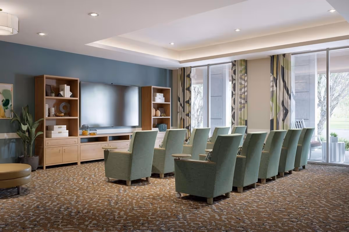 A well-lit common lounge with rows of green upholstered chairs facing a large wall-mounted TV and wooden shelving, with windows and patterned curtains.