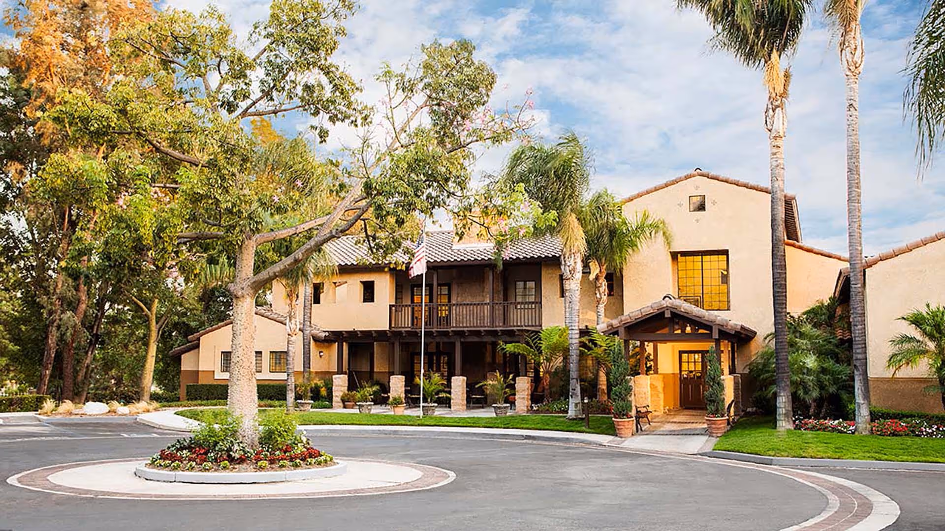 Exterior view of Atria Del Rey senior living facility showing a two-story building with a tiled roof, surrounded by palm trees and other greenery. There is a circular driveway with a landscaped center island featuring a tree and flowers. The entrance has a covered porch with potted plants and benches.