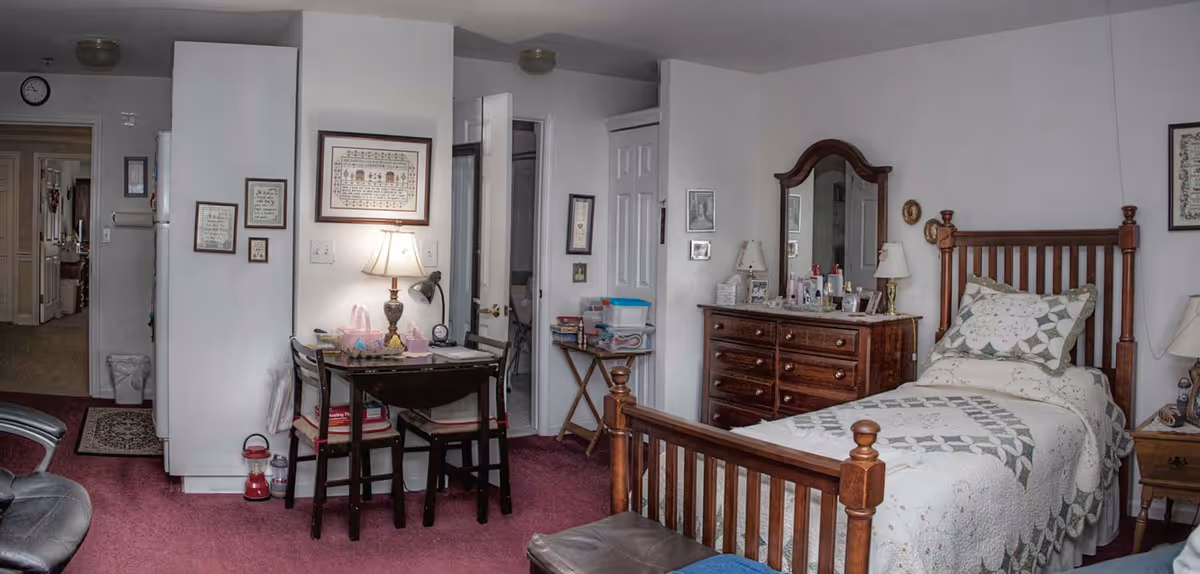 A cozy bedroom in an assisted living facility featuring a wooden bed with a quilted bedspread, a wooden dresser with a mirror, a small table with two chairs and a lamp, and various framed pictures on the walls. The room has a carpeted floor and a door leading to another room or hallway.