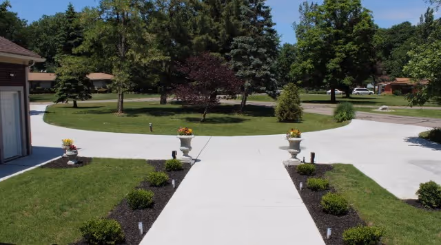 A paved walkway leading to a circular driveway surrounded by green grass, small bushes, and trees under a clear blue sky. There are two decorative flower pots with orange flowers on either side of the walkway near the driveway.