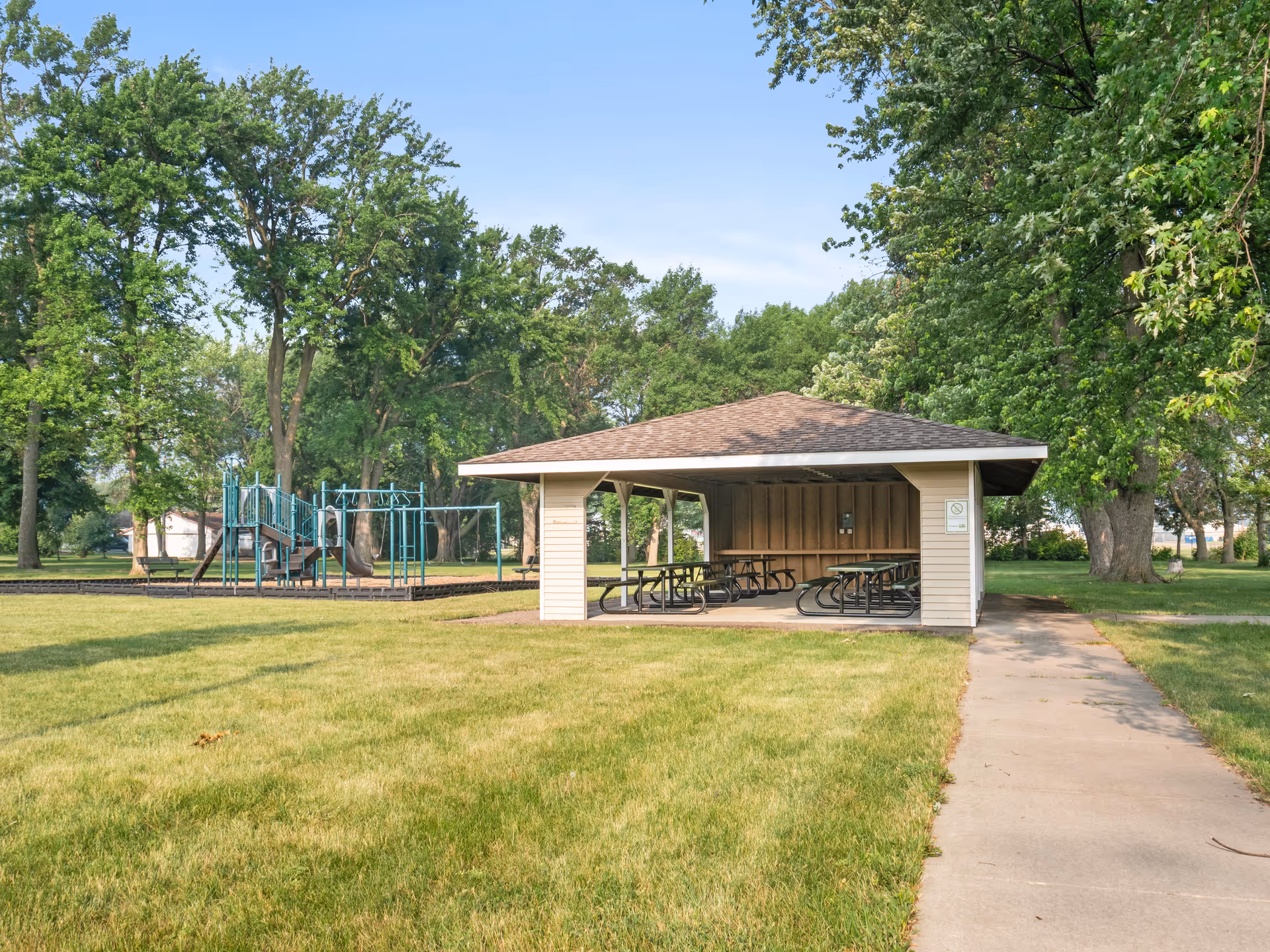 A covered outdoor pavilion with picnic tables on a concrete slab, surrounded by green grass and large trees. To the left, there is a playground structure with slides and climbing equipment. The sky is clear and blue.